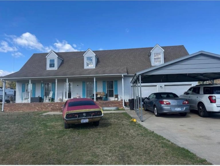 White single-story house with brick foundation, dormer windows, front porch, carport with parked vehicles, and red car in driveway