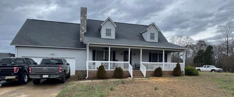 Two-story white house with porch and attached garage, parked vehicles in driveway, bare yard with trees